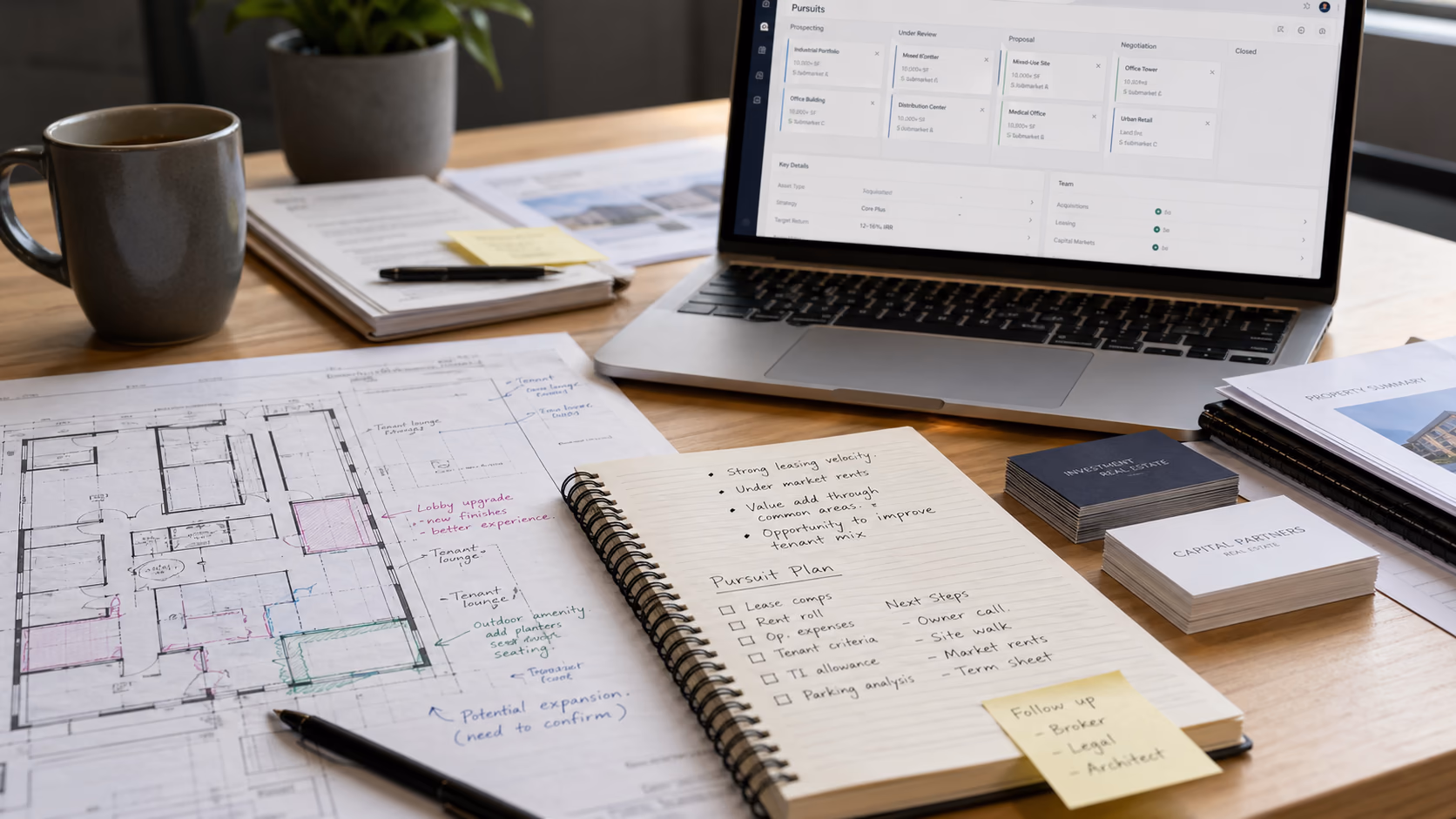 A close view of commercial property notes, marked-up plans, contact cards, and a laptop showing organised pursuit details on a desk