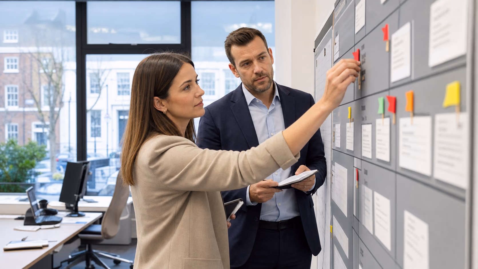 A real estate agent marking exception flags on a shared branch task board while a manager reviews notes beside them in a modern office