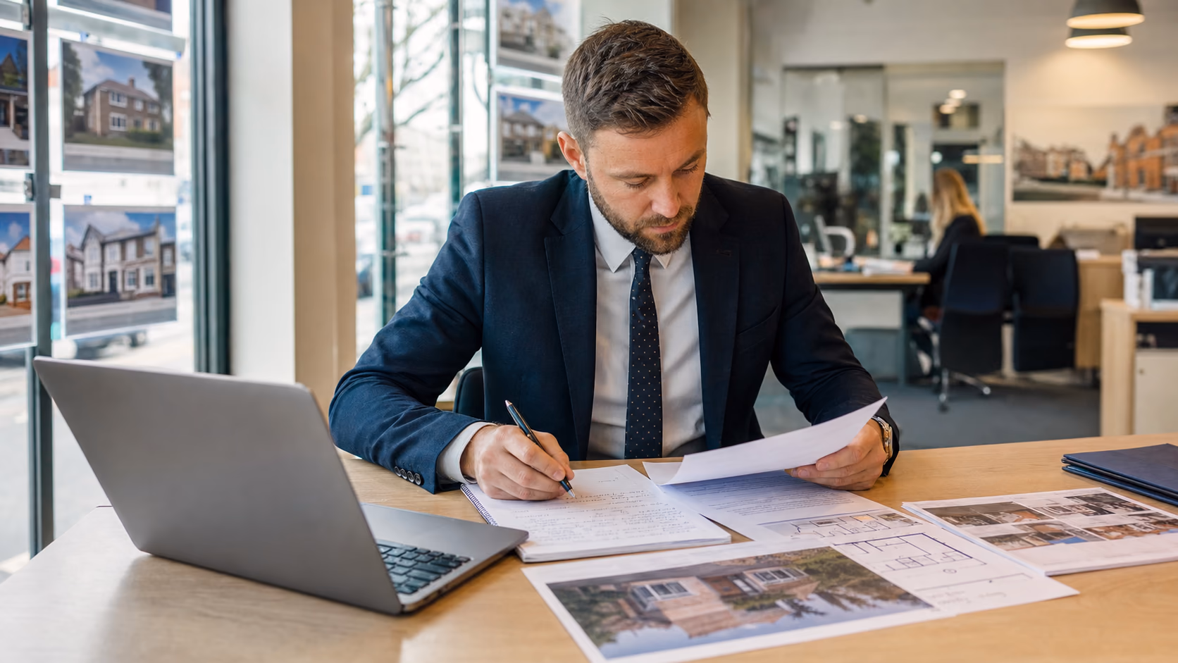 An estate agent reviewing printed listing details, floor plans, and viewing notes beside an open laptop in a busy agency office